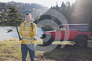 Smiling traveler standing and looking at camera, off-road car and lake in background.