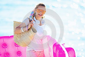 Smiling trendy woman on seacoast holding inflatable mattress