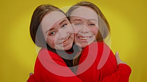 Smiling twin sisters hugging looking at camera posing at yellow background. Portrait of joyful young beautiful slim