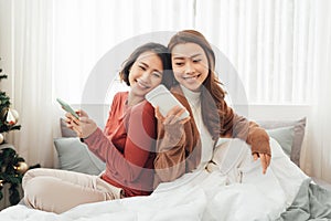 Smiling Two Asian young lovely couple repose on white bed and happy playing with smartphone and social media