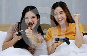 Smiling Two Asian young women lovely couple sitting on white bed and Playing video game with joystick. Funny So glad women