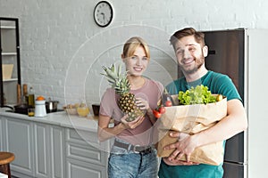 smiling vegan couple with paper bag full of fresh vegetables in kitchen