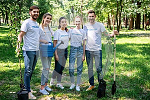 smiling volunteers with new trees watering can and shovel standing in park