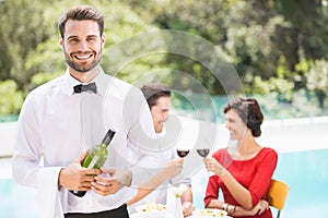Smiling waiter holding wine bottle with couple sitting