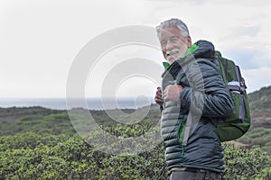 Smiling white-haired elderly man looking at camera while hiking among green bushes and the sea. Horizon over the water. Active