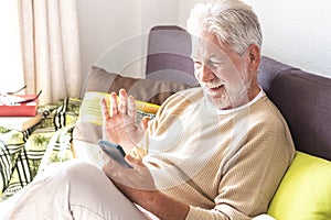 Smiling white-haired senior man relax on the sofa at home video calling with family with his smart phone. Bright light from the