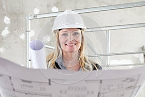 Smiling woman architect or construction engineer reading blueprint wear helmet inside a building site with scaffolding in the