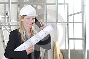 Smiling woman architect or construction engineer talk on the mobile phone wear helmet and holds blueprint inside a building site