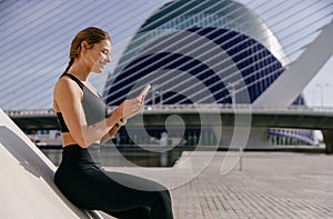 Smiling woman athlete in sportswear sitting outdoors after training and use mobile phone