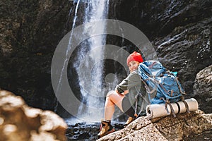 Smiling woman with backpack in red hat dressed in active trekking clothes and boots sitting near mountain river waterfall and