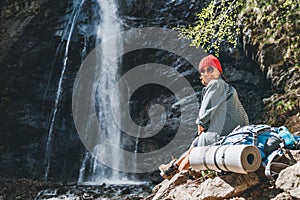 Usmívající se žena s batohem v červeném klobouku oblečená v aktivním trekingovém oblečení a botách sedí u vodopádu horské řeky a