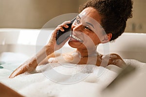Smiling woman, barechested, gestures happily while talking on phone in bathtub