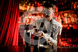 smiling woman bartender holding bottle and pouring drink from it into steel jigger