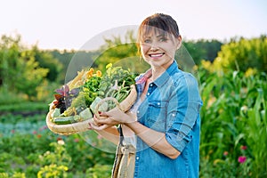 Smiling woman with basket of fresh green vegetables and herbs on farm