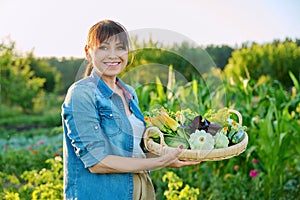 Smiling woman with basket of fresh green vegetables and herbs on farm