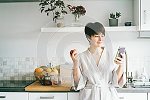 Smiling woman in the bathrobe looking at her mobile phone and eating an apple while standing in the mordern kitchen