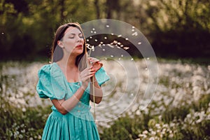 Smiling woman beautiful blowing on ripened dandelion in park. Girl in retro turquoise dress enjoying summer in