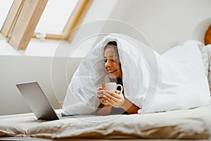 Smiling woman in bed under blanket with coffee and laptop