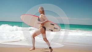 Smiling woman in bikini running with surfboard along sea beach