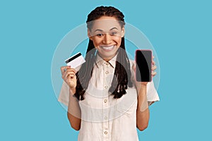 Smiling woman with black dreadlocks showing mobile phone, holding credit bank card, online banking.