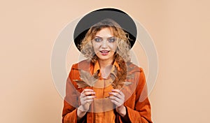 Smiling woman in black hat with autumn leafage. Autumn girl in orange coat with yellow fall leaves. Autumn clothing for