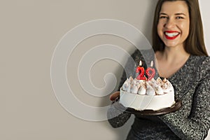 Smiling woman celebrating birthday with cake with number 29 candles on wall background mockup