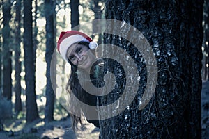 Smiling woman with Christmas hat leaning out behind pine tree in forest