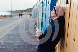Smiling woman with closed eyes in warm clothes with a reusable cup with a hot drink near colorful beach booths and enjoying the