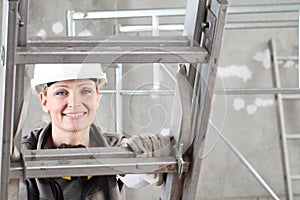 Smiling woman construction worker builder on ladder wearing white helmet and hearing protection headphones on interior site