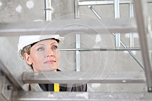 Smiling woman construction worker builder on ladder wearing white helmet and hearing protection headphones on interior site