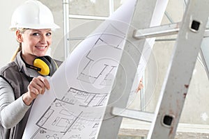 Smiling woman construction worker builder looking at bluprint, wearing helmet and hearing protection headphones in building site