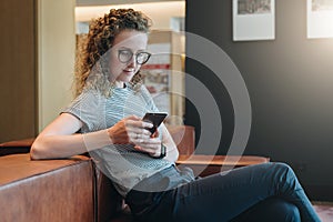 Smiling woman with curly hair in eyeglasses sits in waiting room on couch and uses smartphone. Hipster girl working