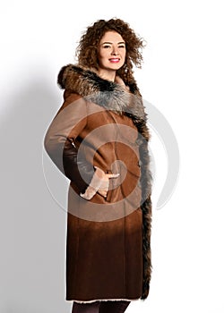 Smiling woman with curly red hair in long brown gradient sheepskin coat with fur collar and cuffs stands in profile