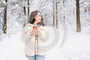 Smiling woman with dark hair in winter clothes with paper cup of coffee in hands against background of snowy winter forest