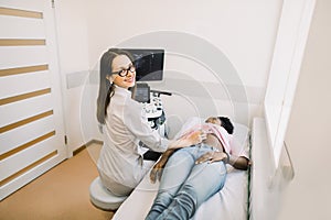 Smiling woman doctor conducting ultrasound examination of patient`s abdomen in clinic. Young African Merican woman