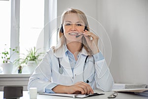 A smiling woman doctor with headphones and a microphone in a white coat conducts an online consultation. Ehealth