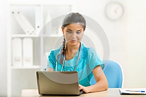 Smiling woman doctor using laptop sitting at desk in medical office