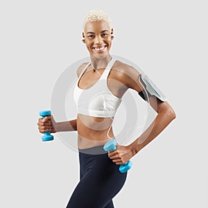 smiling woman doing fitness workout, lifts dumbbells while listening music through earphones of mobile phone on arm band. Latin