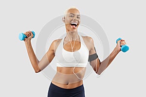 smiling woman doing fitness workout, lifts dumbbells while listening music through earphones of mobile phone on arm band. Latin