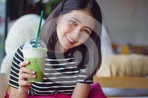 Smiling woman drinking matcha green tea latte in the morning at coffee shop. Portrait pretty Asian girl holding green tea glass in