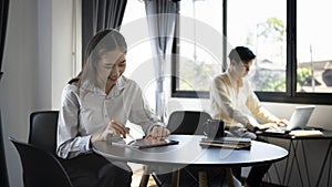 Smiling woman employee working on digital tablet and her colleague sitting in background.