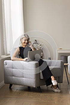 Woman enjoy conversation on smartphone seated on armchair with laptop photo