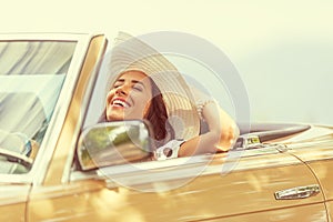 Smiling woman enjoying her summer cabrio ride holding her hat with one hand