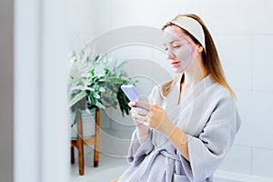 Smiling woman with facial pink clay mask on the face using smartphone at home bath enjoying relaxation and spa beauty