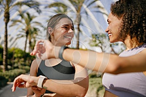 Smiling woman with female friend looking on smartwatch before exercising standing outdoors