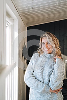 Smiling woman forty year with blonde curly hair in grey knitted sweater near window at house