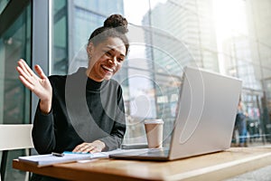 Smiling woman freelance talking with client via video call and waving Hi sitting in cafe