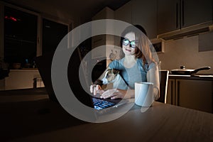 A smiling woman in glasses sits at a wireless computer in the kitchen with a puppy of Jack Russell Terrier on her knees