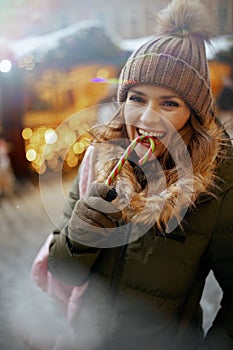 smiling woman in green coat and brown hat at the winter fair in the city with candy cane