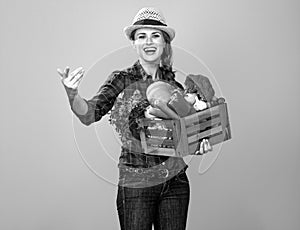 Smiling woman grower with box of fresh vegetables beckoning